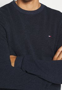 Tummansininen neulepaita, joka on tekstuuripintainen, pyöreäkauluksinen ja jossa on pieni valkoinen, punainen ja sininen logo rinnassa.