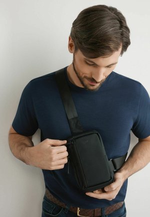 Mężczyzna o krótkich brązowych włosach, ubrany w granatową koszulę, poprawia czarną torbę na pasku na białym tle.
