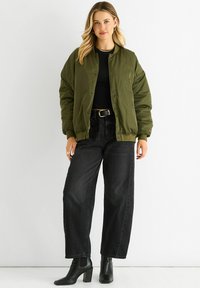 Olive green bomber jacket with a smooth, matte finish, snap buttons, and ribbed cuffs, paired with high-waisted black jeans and ankle boots.
