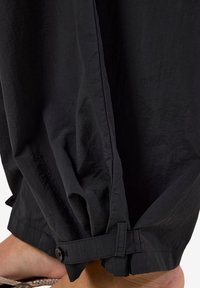 Pieds nus portant un pantalon noir ample avec des bas ajustables à boutons, debout sur une surface texturée. Le tissu du pantalon semble léger.