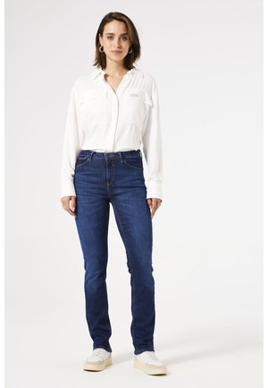 Femme debout portant une chemise blanche à manches longues boutonnée, un jean bleu foncé, des baskets blanches et des boucles d'oreilles créoles dorées sur un fond clair uni.