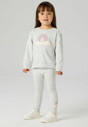 Junges Mädchen steht, trägt ein hellgraues Sweatshirt und Leggings mit pastellfarbenen Regenbogen- und Wolkenmotiven sowie weiße Sneaker.