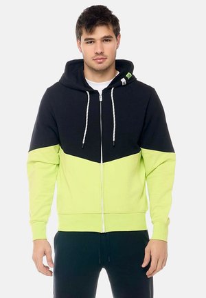 Giovane uomo che indossa una felpa nera e giallo neon con zip e cordoncini, in piedi davanti a uno sfondo grigio chiaro.