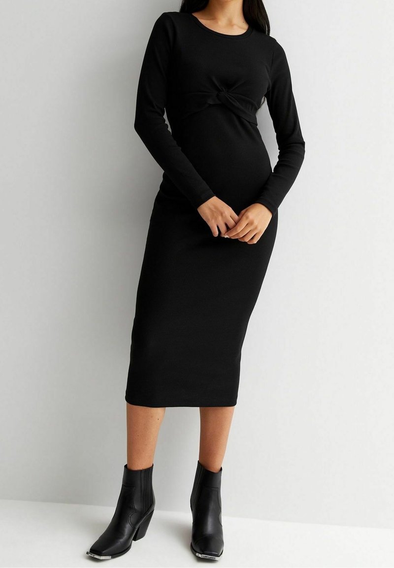 Robe noire à manches longues avec un détail noué à la taille, texture côtelée et silhouette ajustée, associée à des bottines noires à talons.