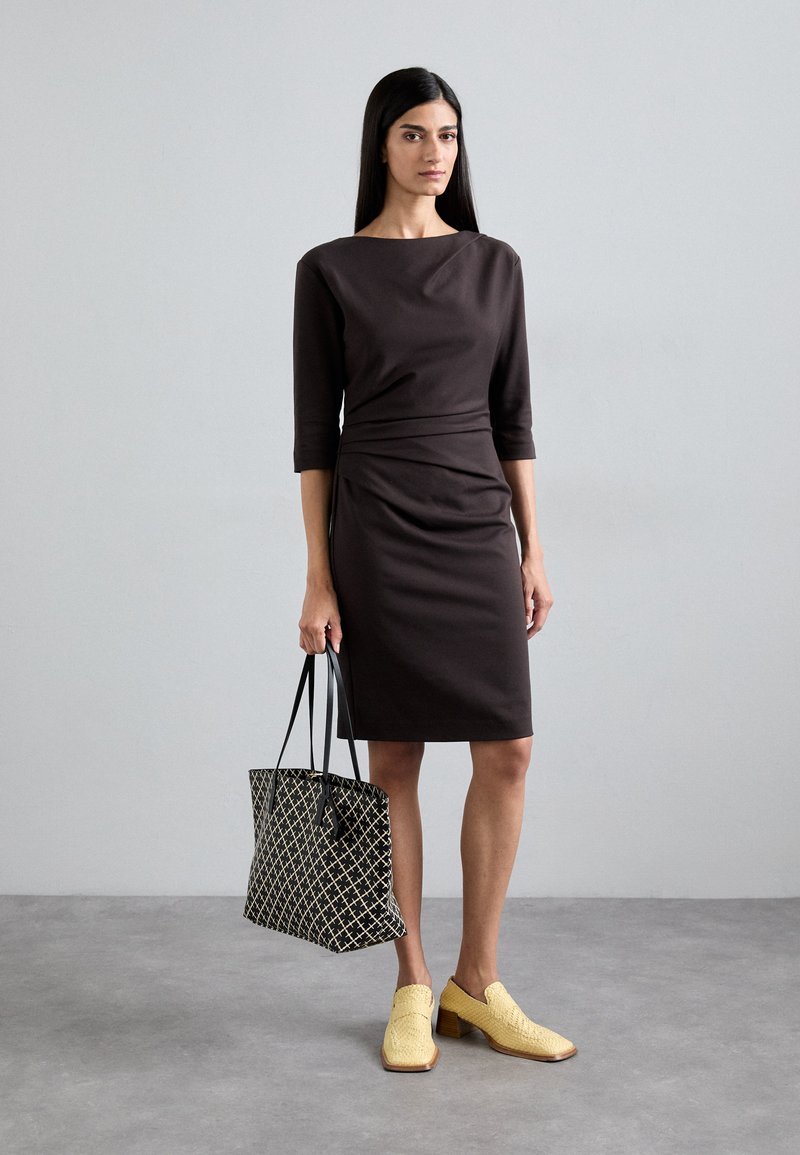 En figurnära brun klänning med trekvartslånga ärmar, texturerad tyg och en samlad midja. Tillbehör inkluderar en svart- och vitmönstrad väska och gula klackskor.