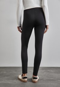 Leggings noires avec une texture lisse et une taille haute, associées à des baskets blanches et noires. L'arrière-plan est gris neutre.