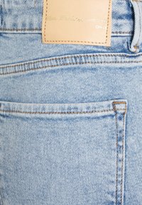 Světle modré džínové kalhoty s koženou nášivkou s embosovaným textem. Tkanina má jemnou texturu a jemné vyblednutí, s viditelným šitím.