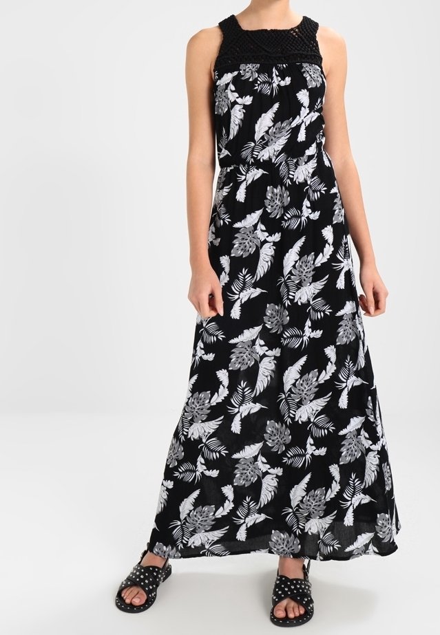Femme portant une robe longue noire sans manches avec un imprimé floral blanc et des sandales noires cloutées, debout devant un fond uni.