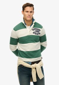 Groen-wit gestreepte rugbyshirt met een kraag en logo-detail op de borst, gecombineerd met donkere jeans en een crèmekleurige kabeltrui om de taille.
