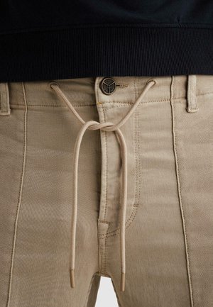 Beige Hosen mit Reißverschluss und Knopfleiste, gesichert durch einen beigen Kordelzug, der in einer Schleife an der Taille gebunden ist, getragen mit einem schwarzen Oberteil.