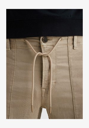 Pantaloni beige con cerniera e patta con bottone, fissati da un cordoncino beige annodato a fiocco in vita, indossati con un top nero.