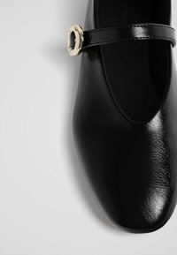 Scarpa piatta nera in pelle con finitura lucida, punta arrotondata e una fascia stretta con dettaglio di fibbia metallica.