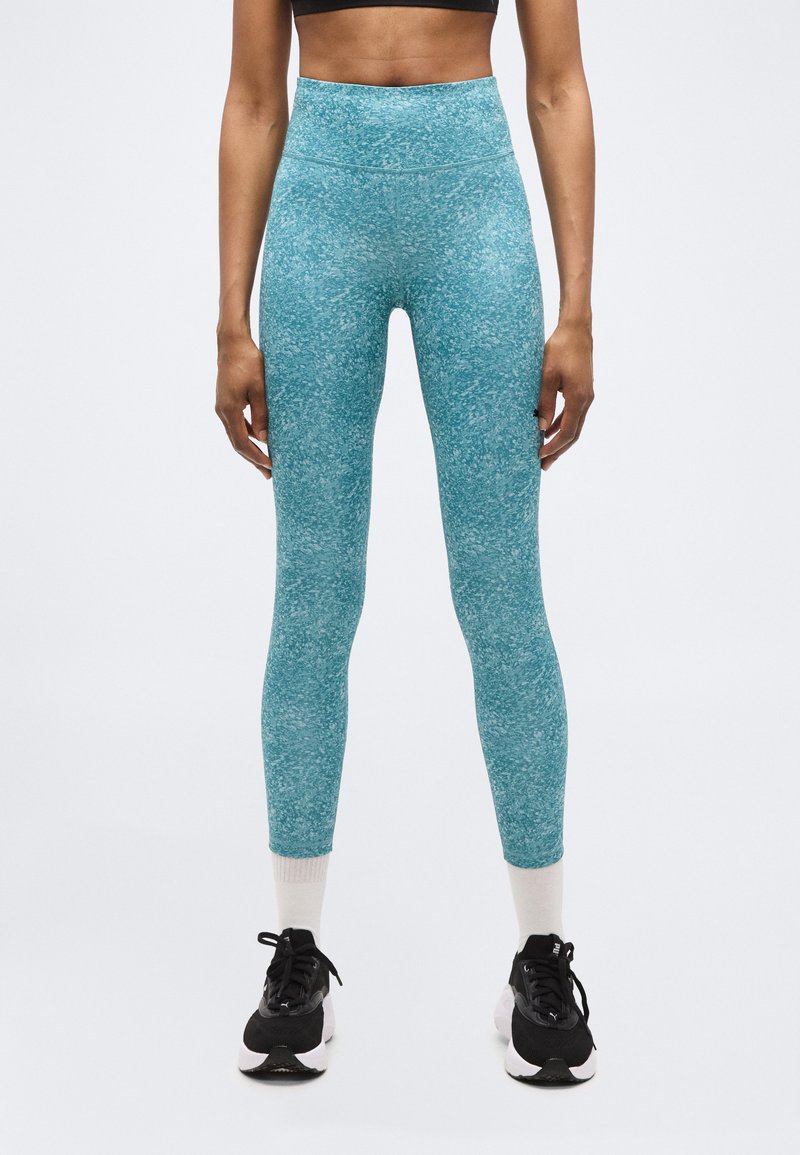 Γυναίκα που φοράει ψηλόμεσα μπλε κολάν με στίγματα, λευκές κάλτσες και μαύρα αθλητικά παπούτσια, στέκεται μπροστά σε απλό λευκό φόντο.