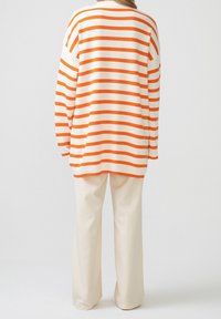 Gestreifter Pullover in Weiß mit orangefarbenen Streifen, mit lockerem Schnitt und weiten Ärmeln, kombiniert mit cremefarbenen, weiten Hosenbeinen.