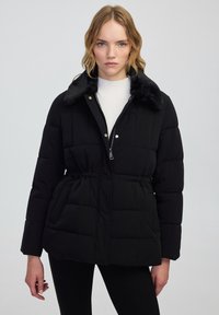 Zwarte pufferjack met een ritsluiting en een kraag van nepbont. Met een getailleerde taille, heeft het een gewatteerde textuur en zijzakken.