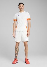 Bela atletska majica s oranžnimi akcenti in modrim logotipom, v kombinaciji z belimi kratkimi hlačami. Oblečena z oranžnimi športnimi čevlji. Ima tetovažo na nogi.