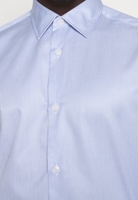 Světle modrá pruhovaná košile z bavlny s límečkem na knoflíčky a bílými plastovými knoflíky. Hladká textilie s jemnými svislými liniemi.