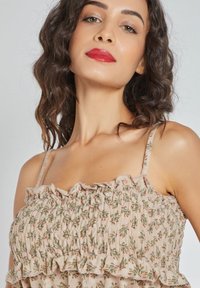 Mujer con cabello castaño ondulado lleva una blusa rosa con flores y volantes, y pintalabios rojo, posando frente a un fondo gris claro liso.