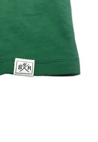 Grøn bomuld T-shirt med ribbet tekstur, der har en hvid etiket med sort tekst og krydsede økser ved kanten som branding.