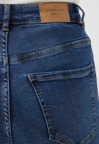 Jeans de denim azul-escuro com costura amarela, apresentando um rótulo retangular de couro castanho com a inscrição "perfect jeans" na parte traseira da cintura.