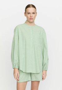 Cămașă verde în dungi cu mâneci lungi, croi lejer, și buzunar pe piept, purtată împreună cu pantaloni scurți asortați cu același model.