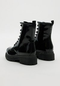 Botas de combate negras, brillantes, con cordones, suelas gruesas y tiradores en la parte posterior, mostradas sobre un fondo claro y sencillo.