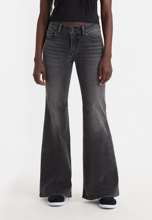 Personne portant un jean flare gris foncé taille haute, associé à des baskets noires et un crop top noir, debout devant un fond blanc.
