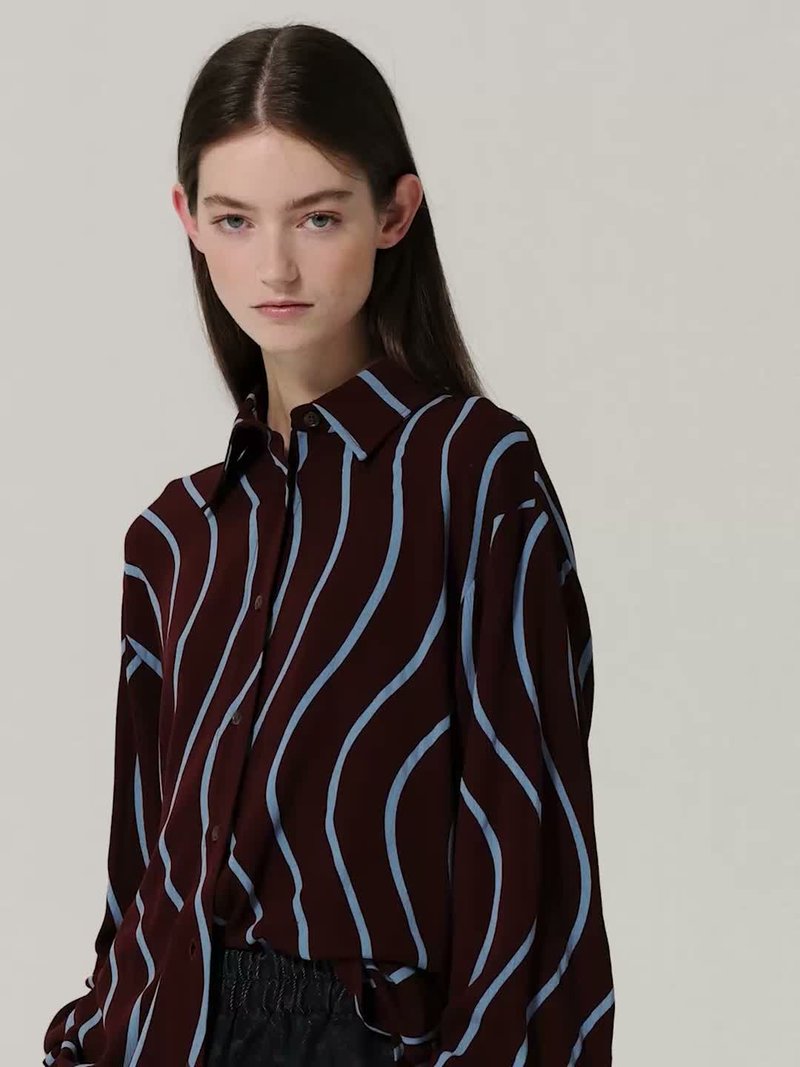 Jeune femme aux longs cheveux foncés portant une chemise sombre avec des rayures verticales ondulées bleu clair sur un fond clair uni.