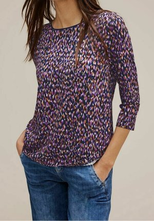 Femme portant un haut à manches longues aux motifs violets, noirs et orange avec un jean bleu, mains dans les poches, sur un fond neutre.