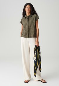Olijfgroene kortemouwen shirt met een textuur van bladeren, gecombineerd met witte wijde broekspijpen en een gedessineerde sjaal die in één hand wordt vastgehouden.