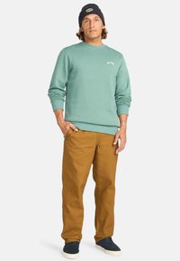 Felpa verde menta con polsini a coste, abbinata a pantaloni in tela marrone e scarpe blu navy. Modello con un berretto blu navy abbinato.