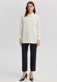 Krémová blůza s límečkem, předním zapínáním na knoflíky a dekoračním lemováním na ramenou, spárovaná s černými slim-fit kalhotami a černými lodičkami.