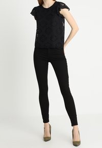 Top de encaje negro con mangas cortas y volantes, patrón floral, combinado con pantalones skinny negros y tacones altos verde olivo.