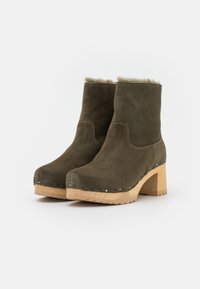 Botas de tobillo de ante verde con un ribete de piel sintética, tacón de madera y costuras blancas a lo largo de la suela. Diseño de tacón bloqueado con punta redonda.