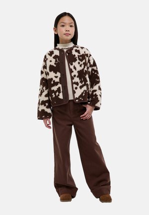Jeune personne debout portant un col roulé blanc, une veste boutonnée marron et blanche à motifs, un pantalon large marron et des chaussures marron.