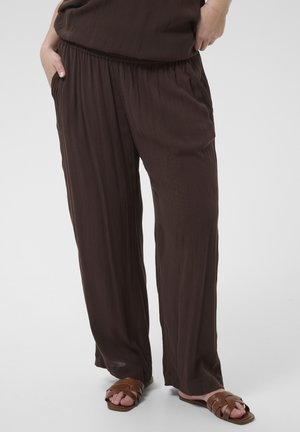 Pantalon ample marron foncé porté par une personne les mains dans les poches, associé à des sandales plates tressées marron sur fond blanc.