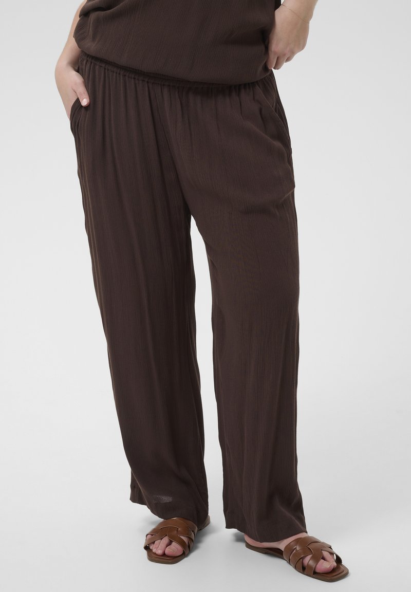 Pantalon ample marron foncé porté par une personne les mains dans les poches, associé à des sandales plates tressées marron sur fond blanc.