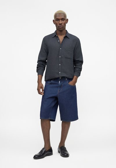 Μαύρος άνδρας που στέκεται με το χέρι στην τσέπη του, φοράει σκούρο πουκάμισο με μακριά μανίκια και κουμπιά, μπλε τζιν σορτς και μαύρα δερμάτινα μοκασίνια.