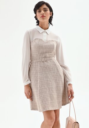 Donna che indossa un vestito beige testurizzato con cintura sopra una camicia bianca a maniche lunghe con colletto, tenendo una borsa beige al fianco.