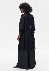 Czarny długi płaszcz z koronki o luźnym kroju, marszczonych rękawach i lekko zaokrąglonym brzegu. Noszony na szerokich czarnych spodniach.