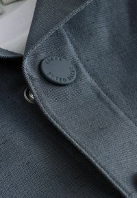 Gros plan sur le revers texturé gris foncé d'un blazer avec un bouton mat rond embossé de la mention "Baker by Ted Baker".