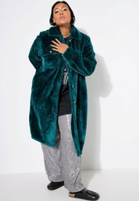 Cappotto in pelliccia sintetica color turchese con colletto e chiusura a bottoni, indossato sopra una maglietta nera e pantaloni di paillettes argentati. Mocassini neri.