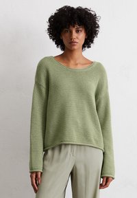 Grüner Strickpullover mit lockerer Passform, weitem Bootsausschnitt und langen Ärmeln. Strukturiertes Muster und gerippter Saum. Kombiniert mit hellen Hosen.