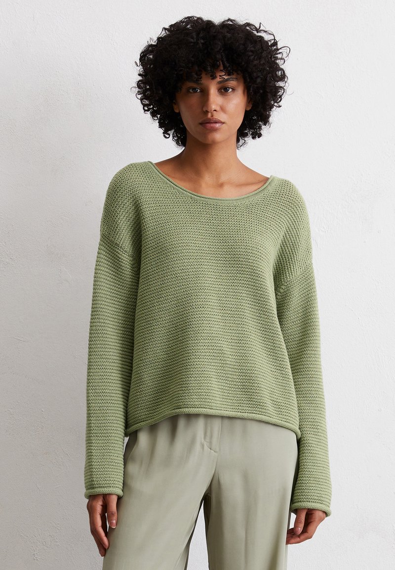 Grüner Strickpullover mit lockerer Passform, weitem Bootsausschnitt und langen Ärmeln. Strukturiertes Muster und gerippter Saum. Kombiniert mit hellen Hosen.