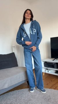 Sudadera con capucha azul con cremallera y logo GAP reflectante, camiseta gris claro, vaqueros azules y zapatillas con estampado; de pie sobre una alfombra gris claro en una habitación.