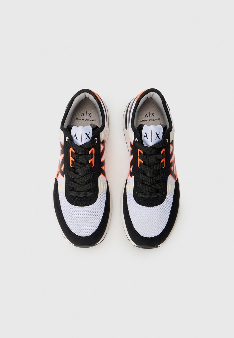 Zapatillas deportivas en blanco y negro con acentos naranjas, parte superior de malla, lengüeta acolchada y suela blanca. Presenta el logotipo en la plantilla y la lengüeta.