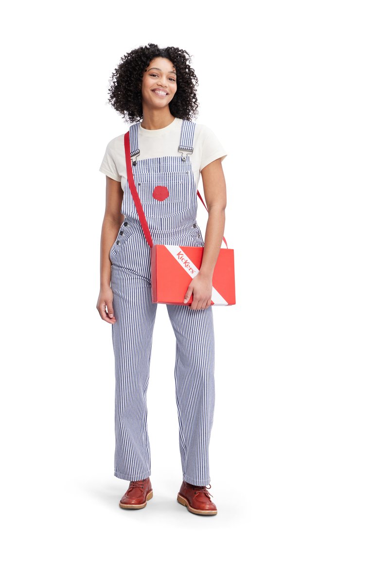 Salopette a righe blu con dettaglio a cerchio rosso, abbinata a una t-shirt color crema, una borsa a tracolla rossa e scarpe marroni.