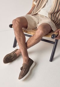 Brown suede loafers with decorative woven pattern and tassel detail, paired with beige shorts, seated on a woven chair.