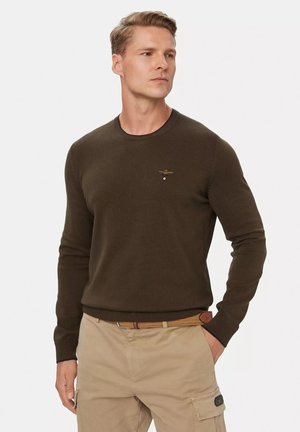Uomo che indossa un maglione girocollo verde scuro e pantaloni cargo beige, in piedi con una mano in tasca, guardando leggermente di lato.