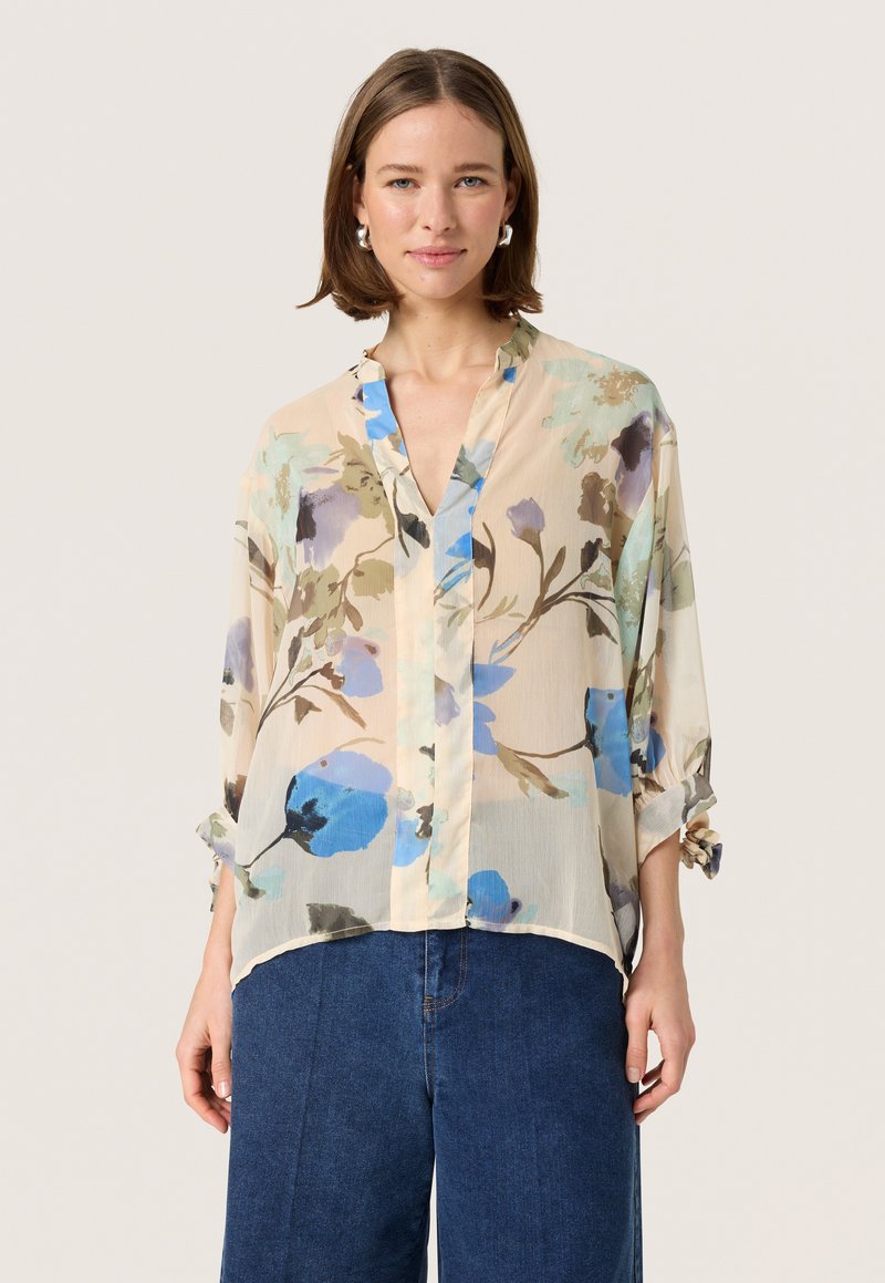 Blusa floreale in un tessuto leggero e trasparente con una base beige, caratterizzata da fiori blu e verdi, scollatura a V e maniche arrotolabili.